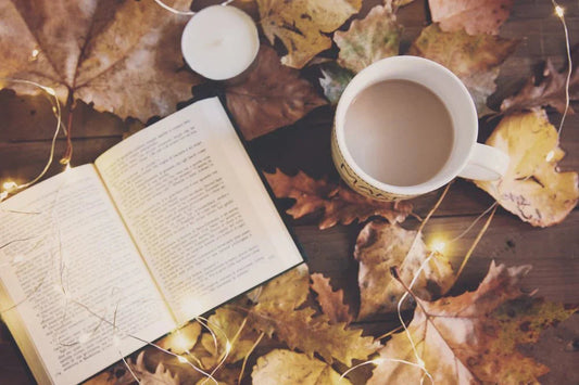 Open book and coffee on wooden table with autumn leaves, fairy lights, and candle